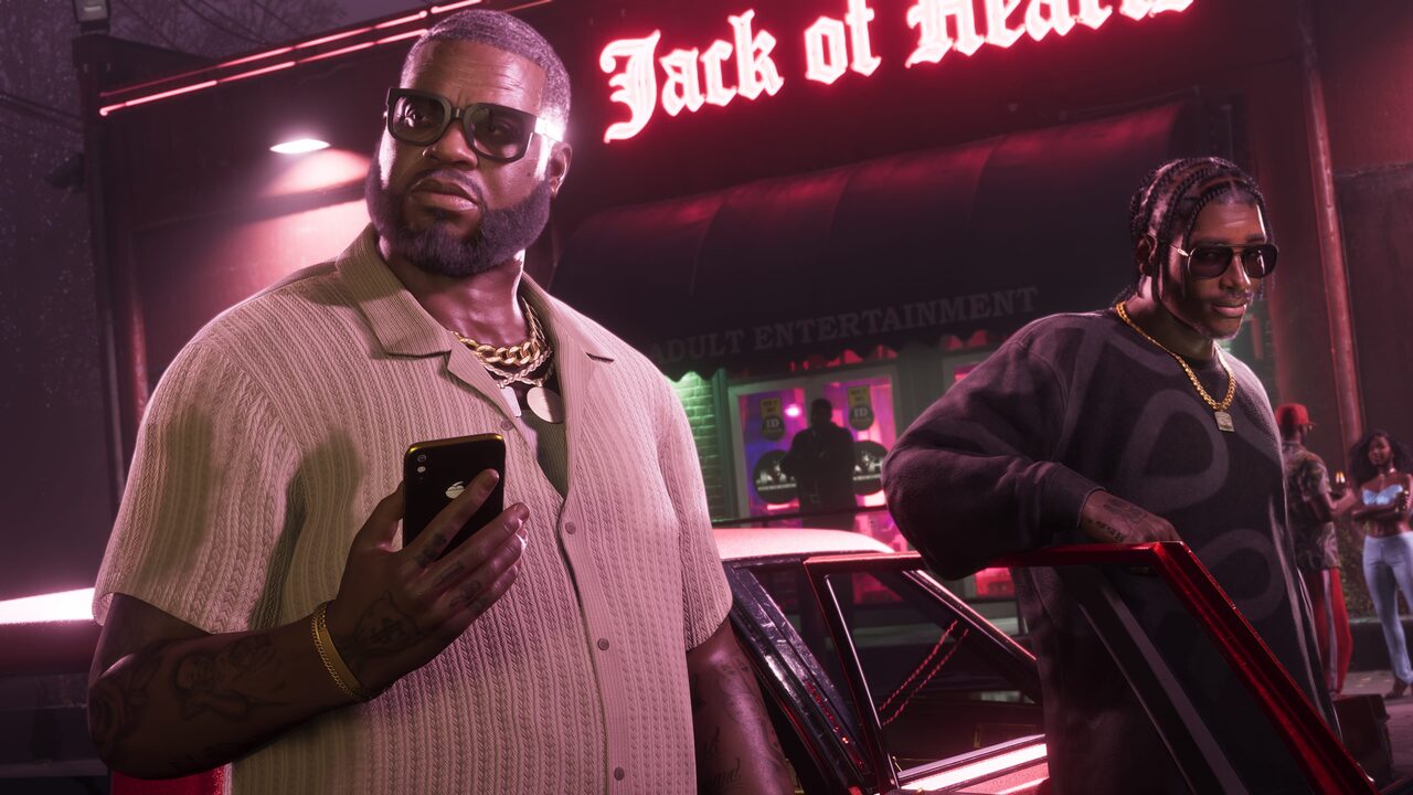 Boobie Ike standing outside the Jack of Hearts club at night holding a phone, another man beside him near a parked car.