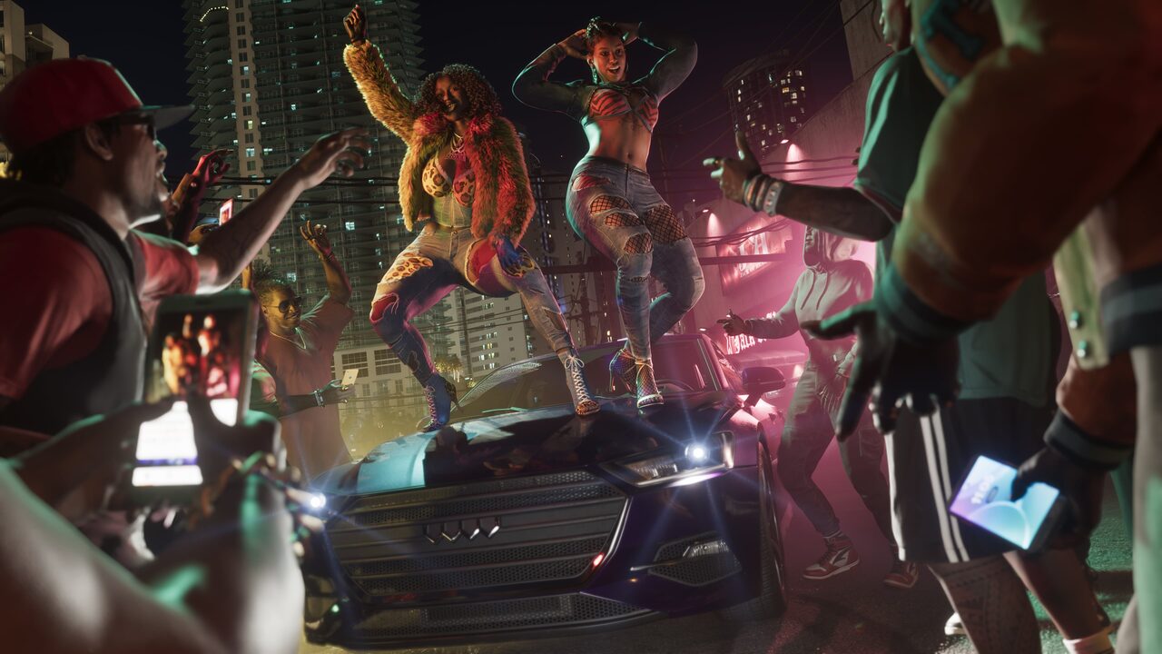 Two women dancing on the hood of a black car at night while the crowd around them films on their phones.