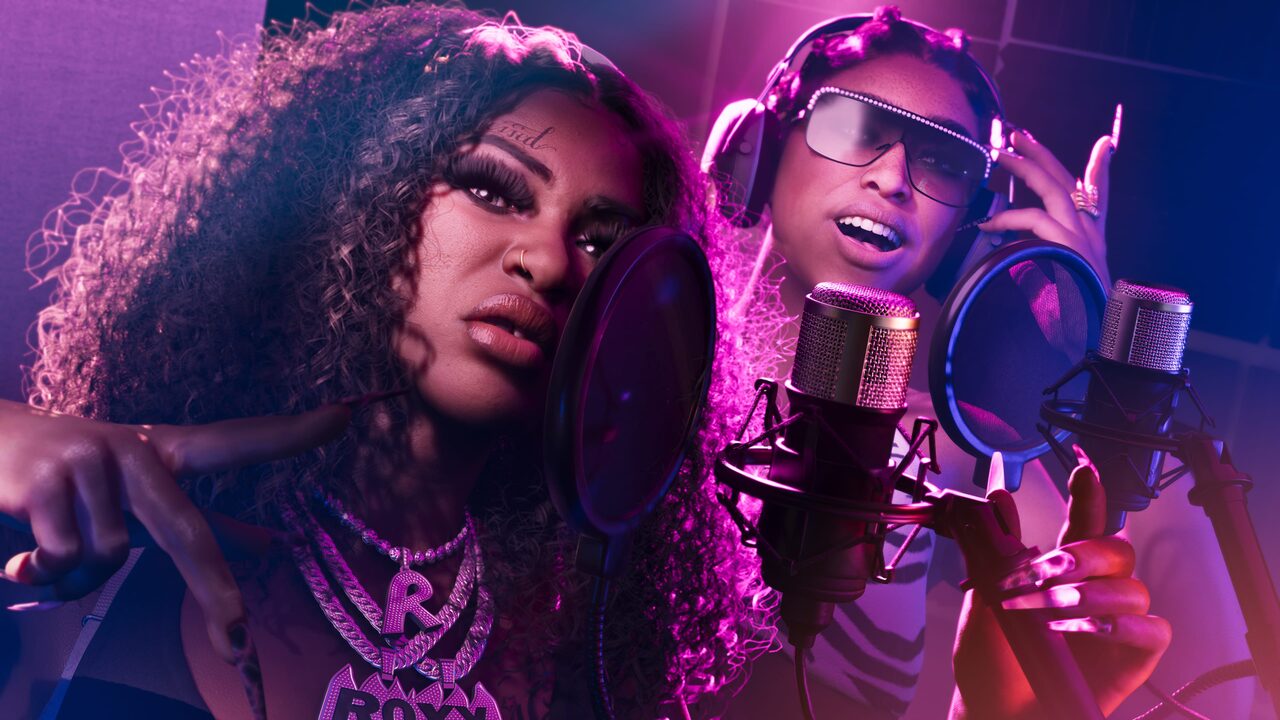 Two women recording in a studio lit pink and purple, one leaning into a microphone and the other in headphones and sunglasses, a ROXY pendant visible.
