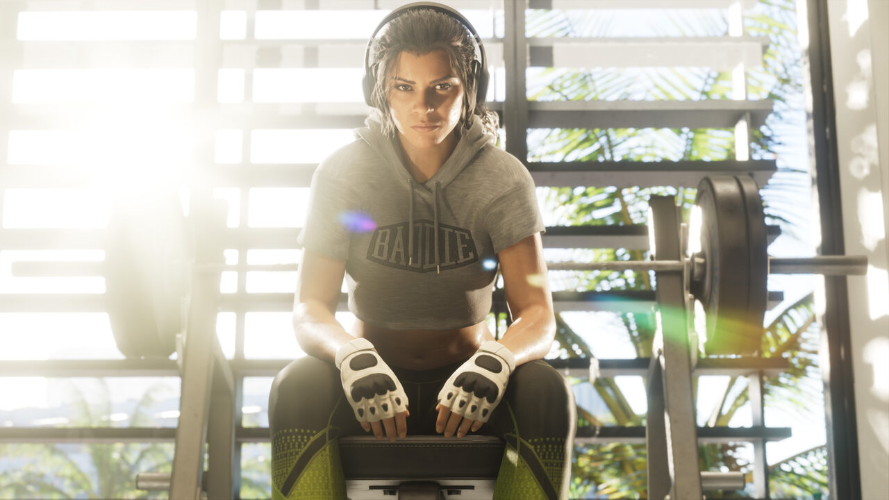 Lucia Caminos sitting on a weight bench in a sunlit gym, wearing headphones, a grey hoodie and fingerless gloves.