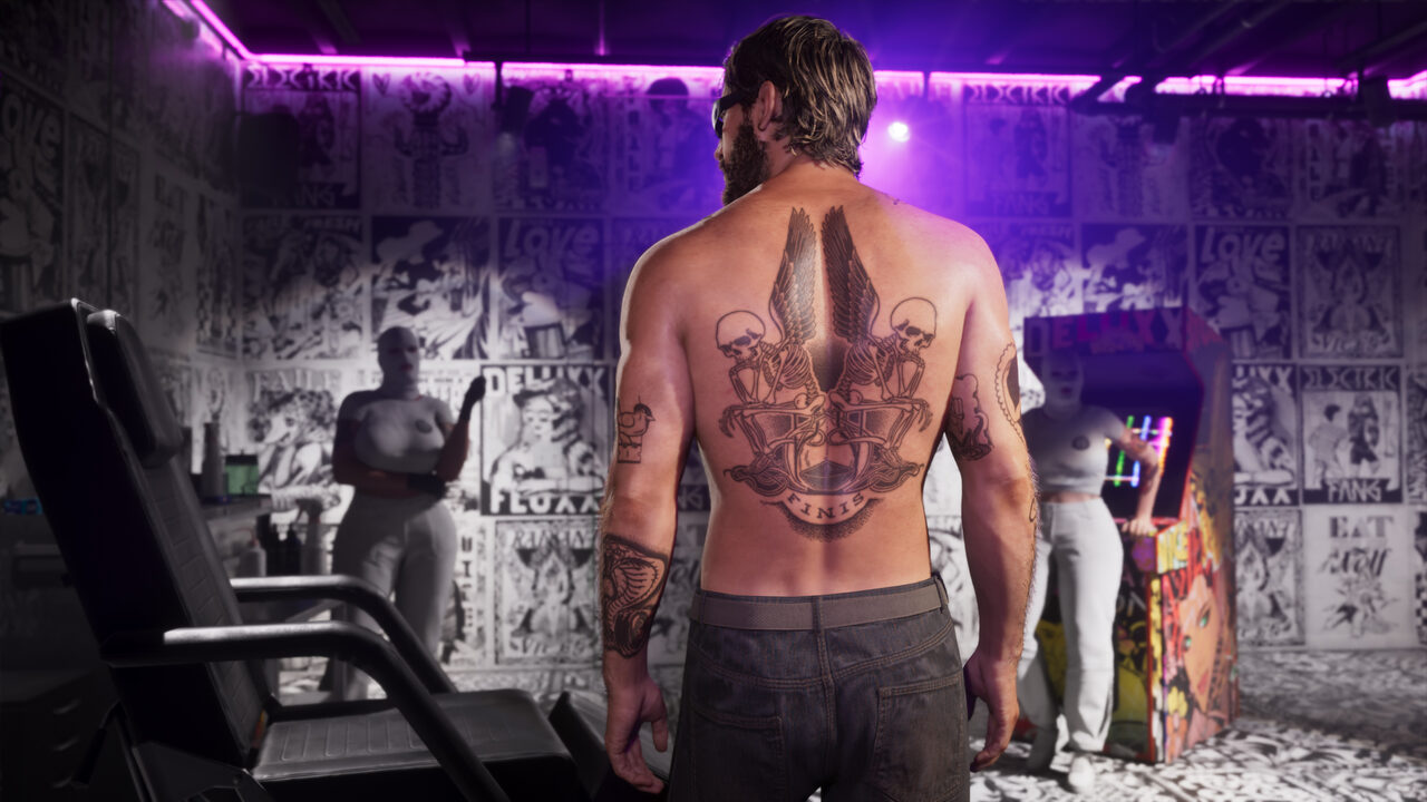 A man seen from behind inside a tattoo studio showing a large winged skeleton back piece, flash-covered walls and an arcade cabinet around him.