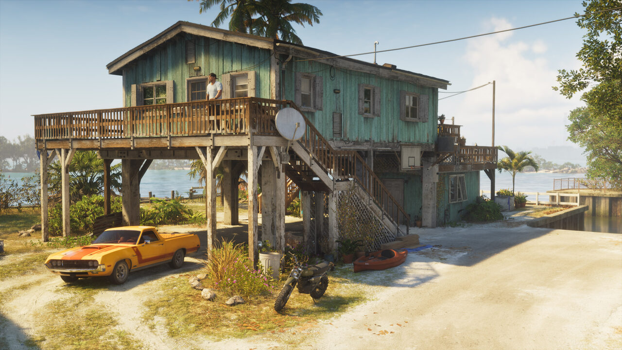 A weathered green stilt house over shallow water, a man on its deck, a yellow coupe utility and a dirt bike parked on the sand beneath.