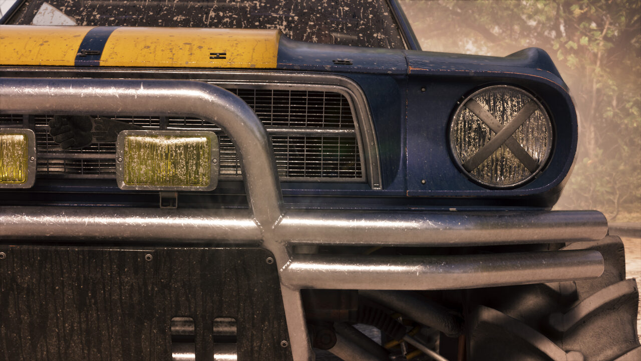 Close front detail of a mud-spattered blue off-road muscle car, its headlight covered by an X-shaped grille guard and yellow driving lamps below.