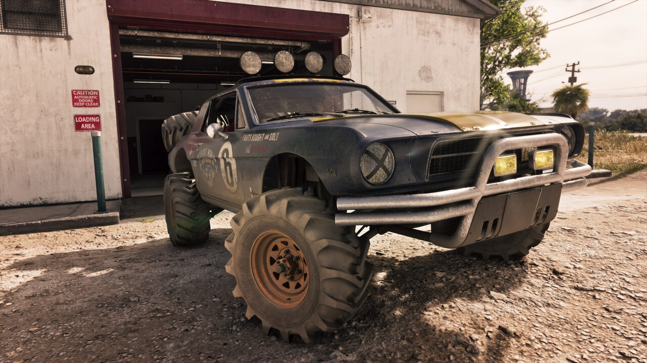 A blue and gold number 6 muscle car lifted on huge off-road tyres, parked on gravel outside a workshop loading door.