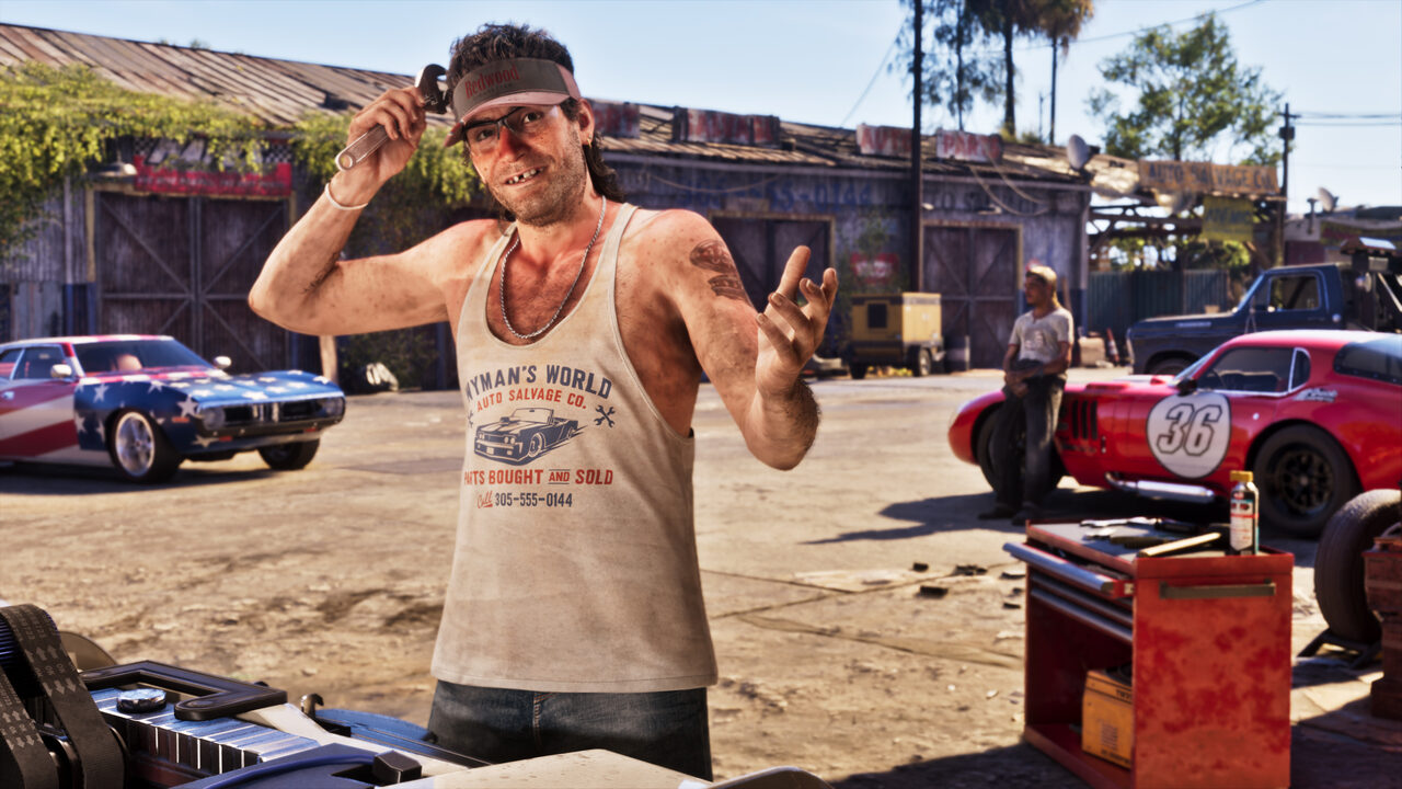 A mechanic in a Wyman's World Auto Salvage vest raising a spanner and grinning in a sunlit salvage yard, a star-spangled muscle car and a red number 36 race car behind him.