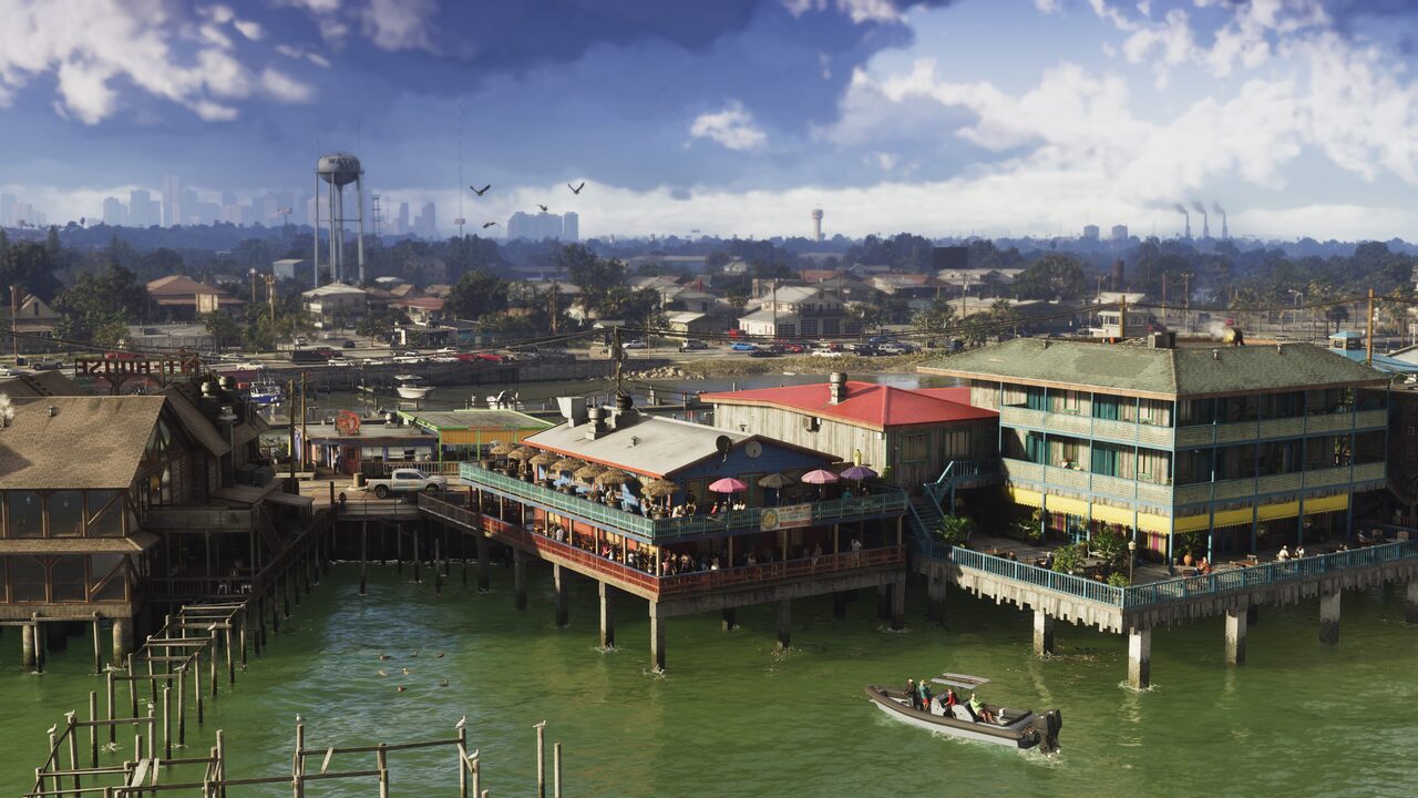 A Grassrivers waterfront built out over green water on pilings, a busy deck restaurant under a red roof beside a teal motel, with a distant city skyline across the bay.