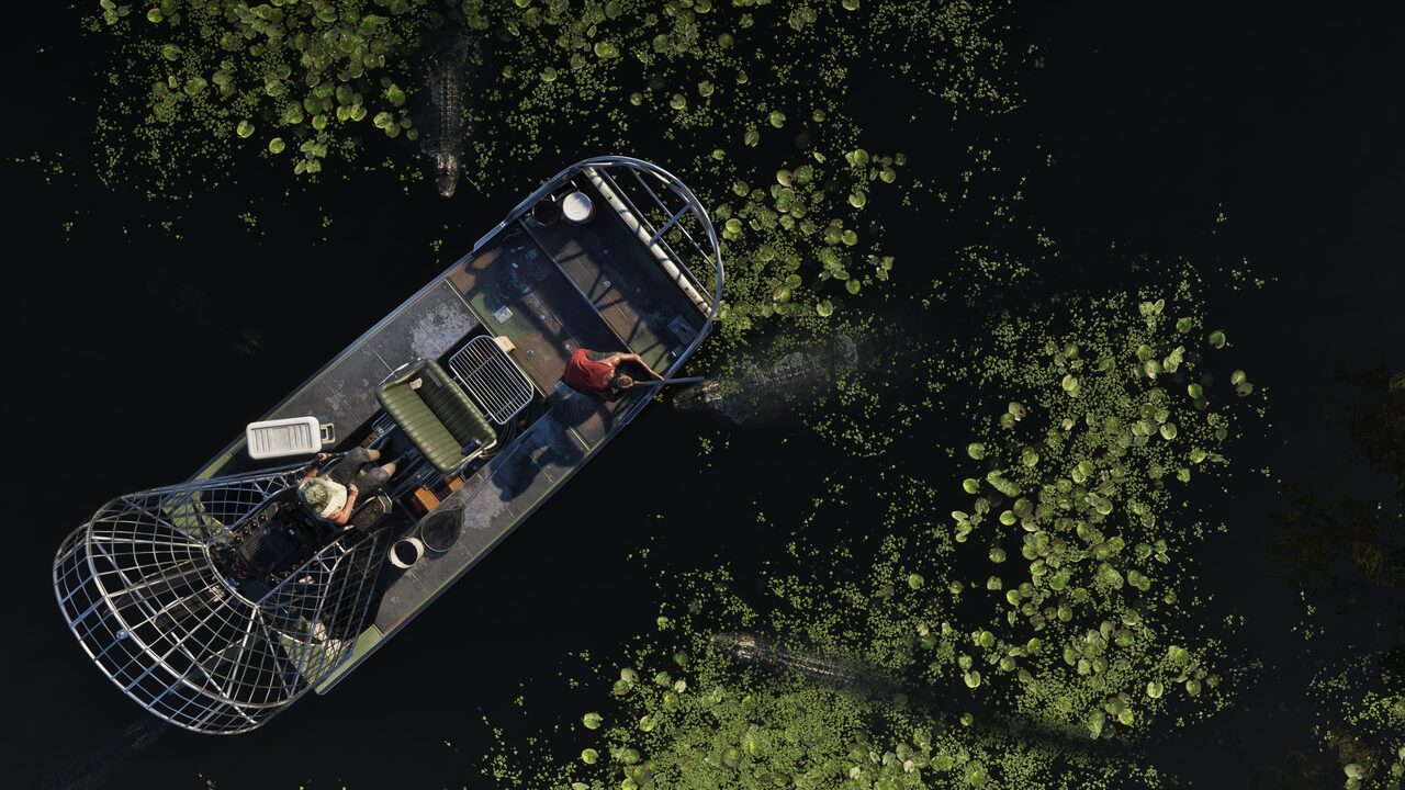 An overhead shot of an airboat idling on black water among lily pads, two people aboard and alligators gliding just beneath the surface.