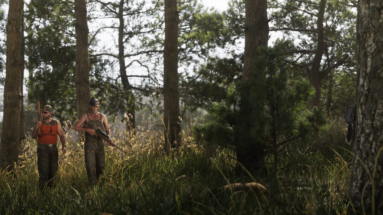 Two men in camouflage carrying rifles through tall grass in a pine wood, one of them in a blaze-orange vest.
