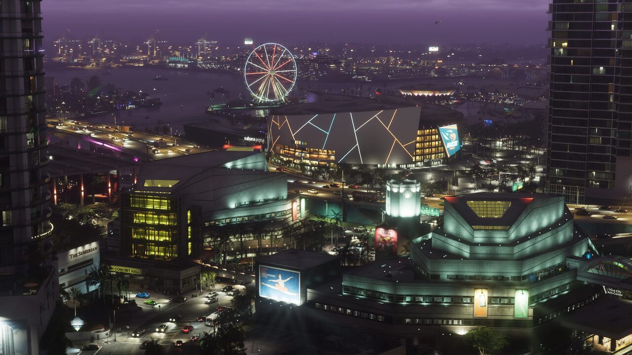 A night aerial over downtown Vice City, an arena, concert hall and arts centre lit below with a Ferris wheel and port cranes on the waterfront behind.